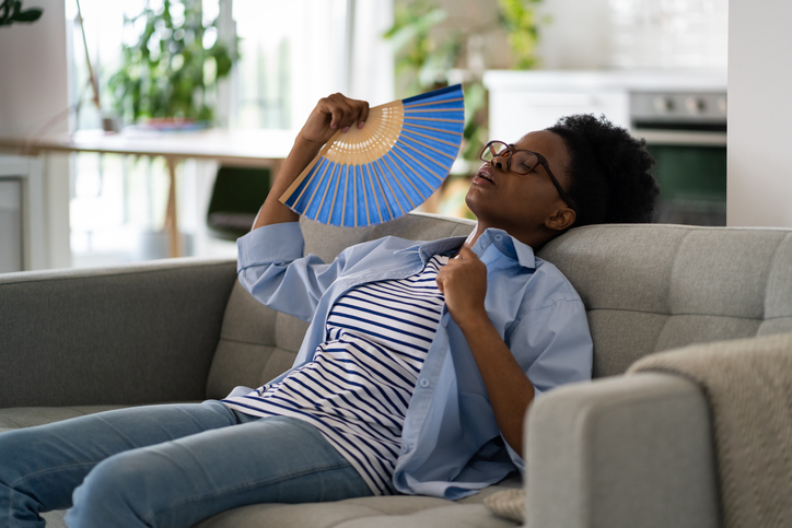 Tired exhausted African American woman sits on sofa at home using paper fans and closing eyes suffering from stuffiness.